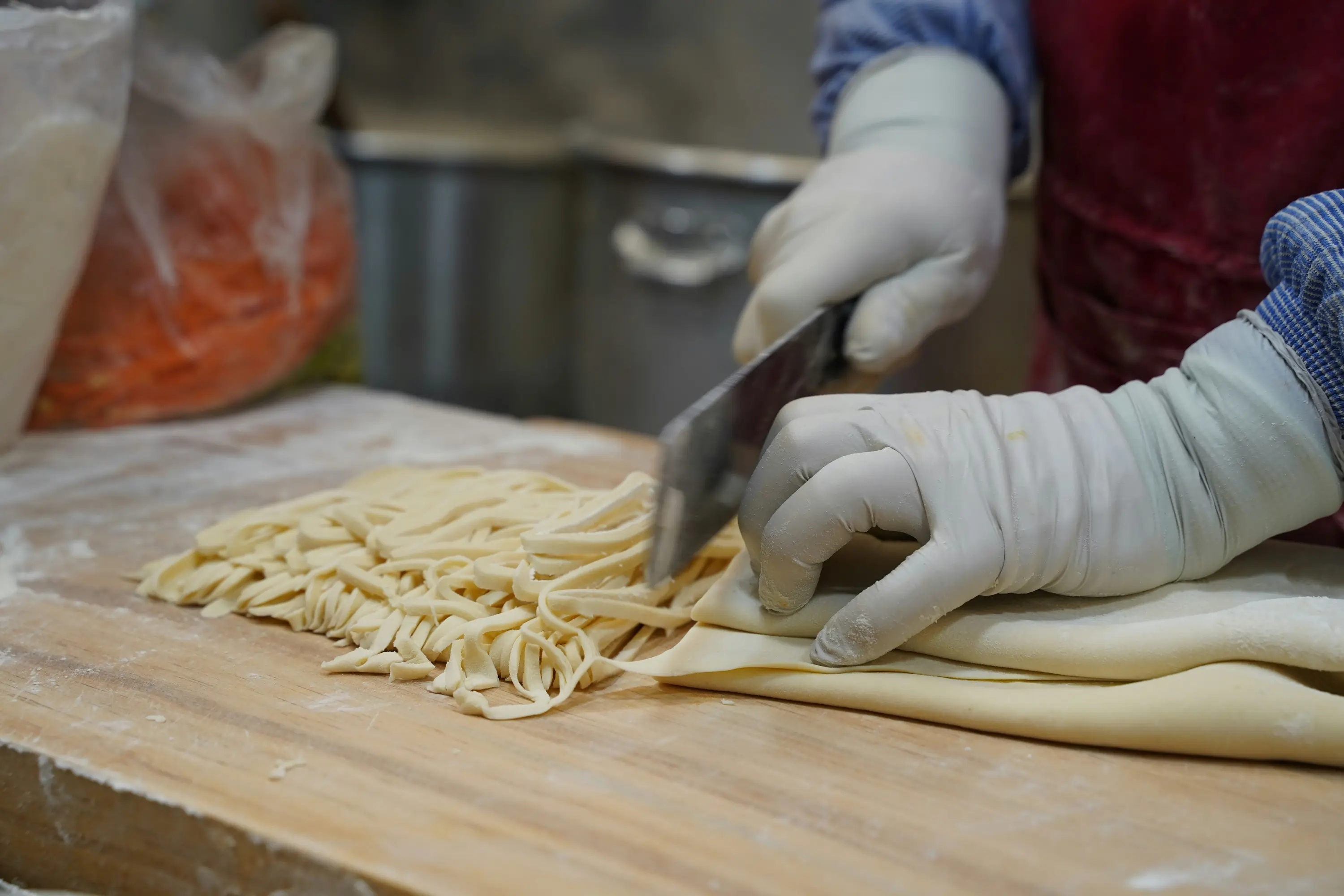 Person wearing gloves cutting handmade dough noodles on a wooden surface.